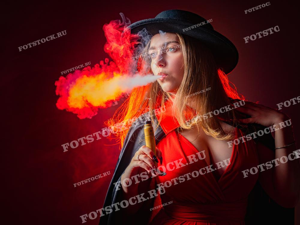 Фотография изображает женщину в черной шляпе и красном платье, выпускающую яркое облако дыма от электронной сигареты.